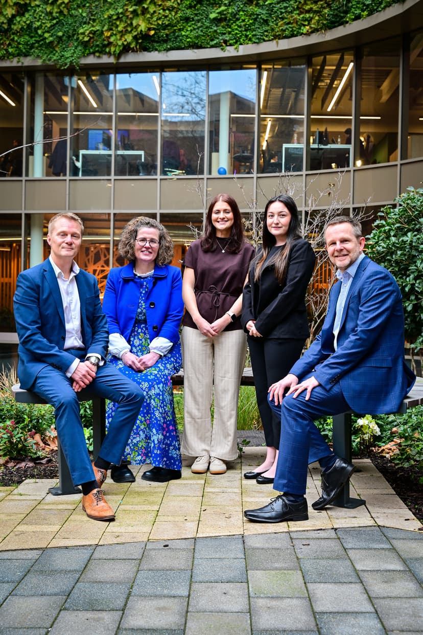 Five professionals pose together in a modern courtyard, surrounded by greenery and contemporary architecture, smiling confidently.