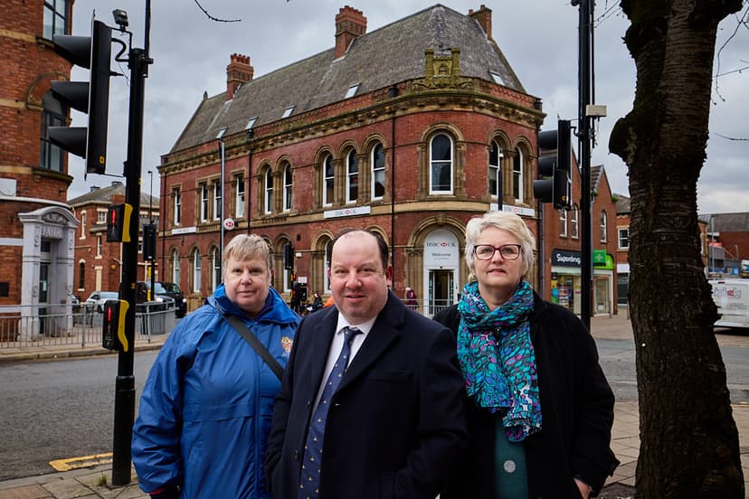 Three individuals stand in front of a historic red-brick building in Wakefield, framed by traffic signals and a cloudy sky.
