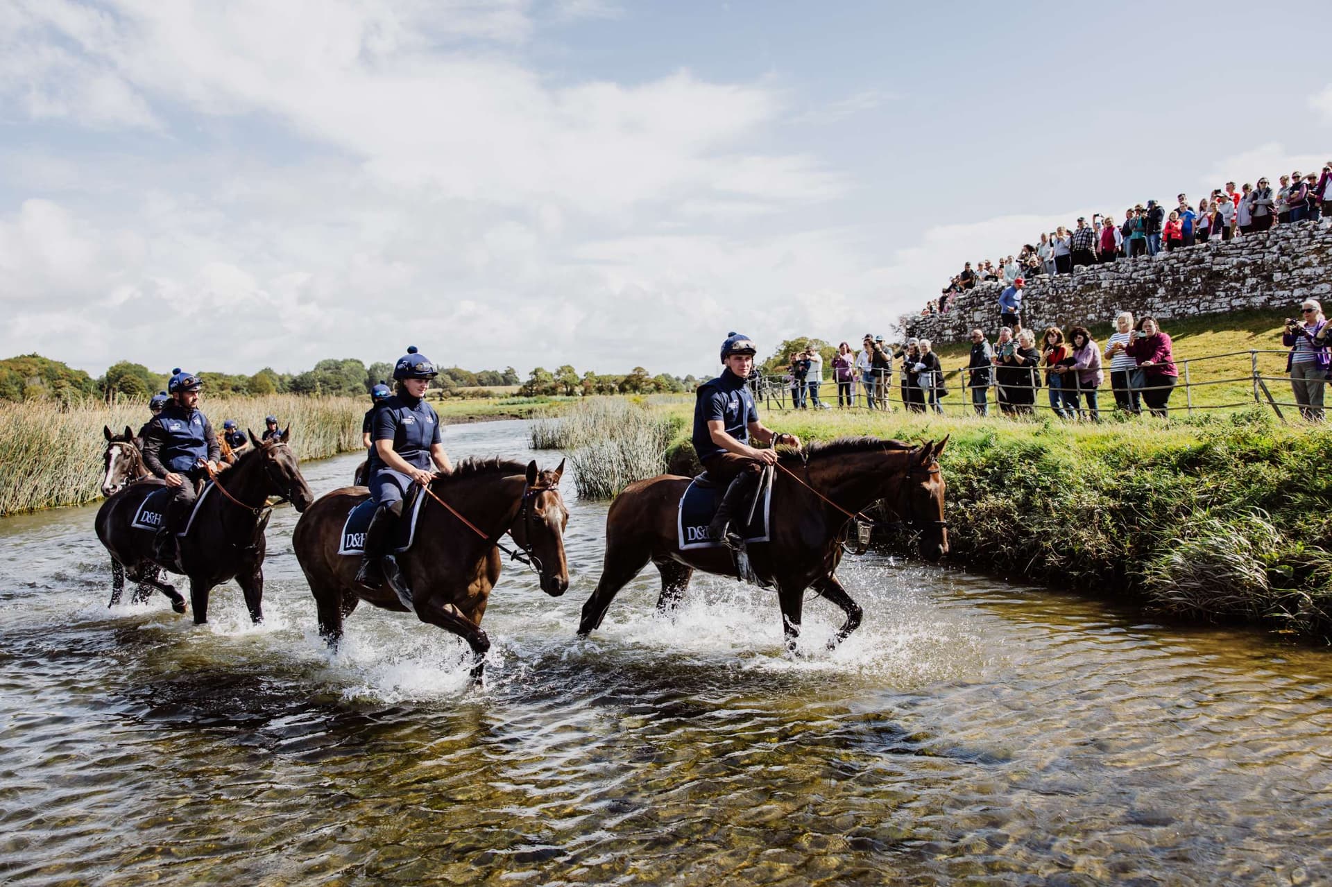 Three riders on horseback crossing a shallow river with spectators on the bank