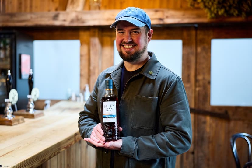 A smiling man in a cap holds a bottle of whisky inside a rustic distillery with wooden beams and bar setup.