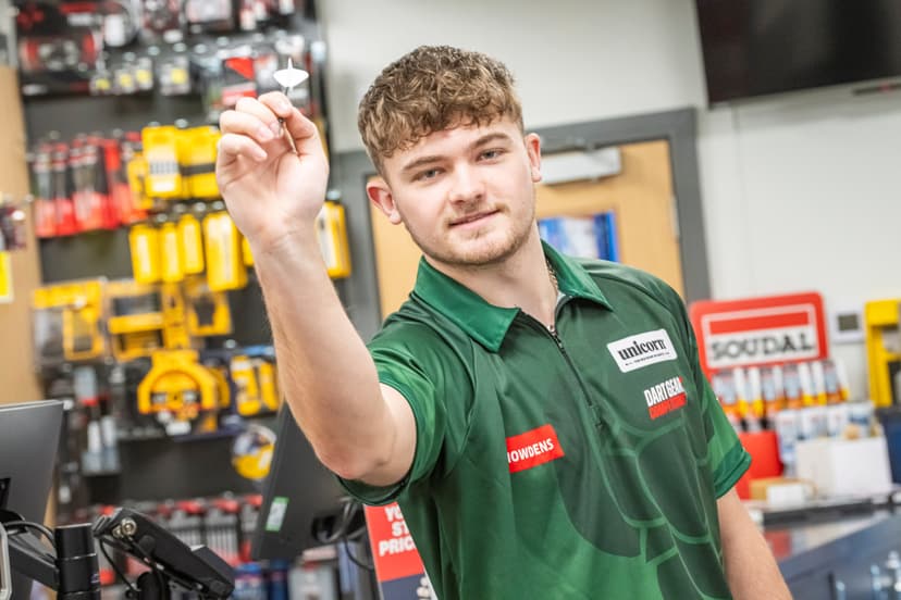 A young man in a green darts shirt holds a dart confidently in a well-stocked hardware store.