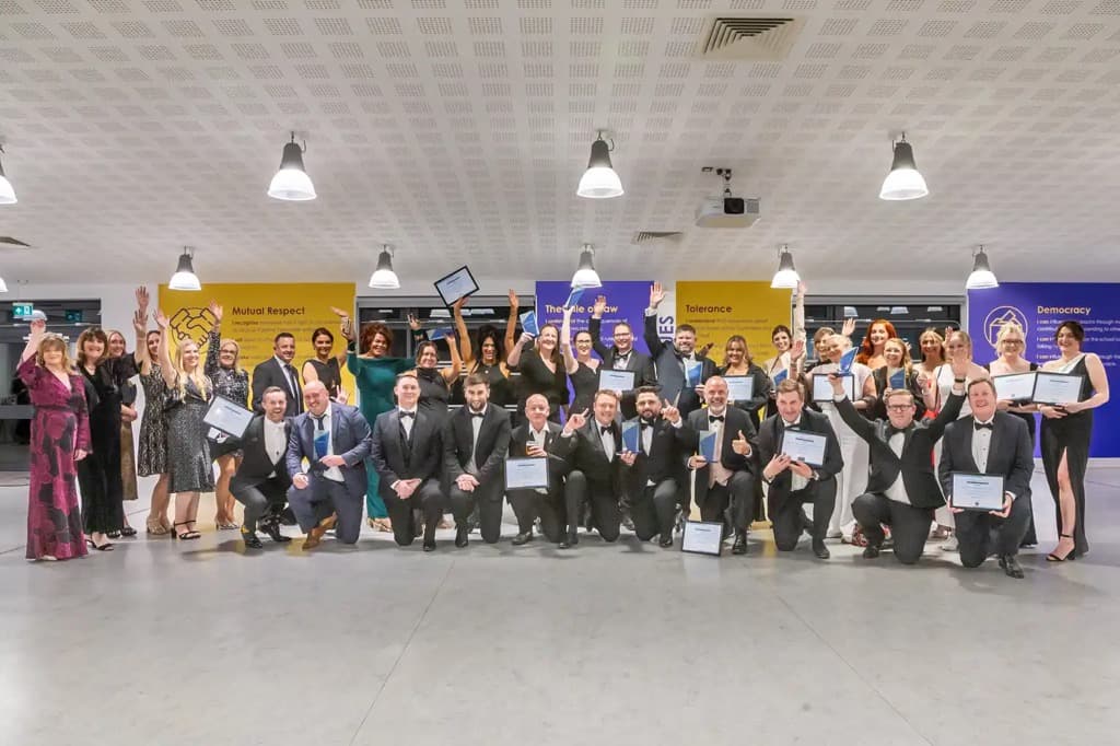 Celebratory group of award winners in formal attire, holding certificates, in a modern venue with colorful banners.