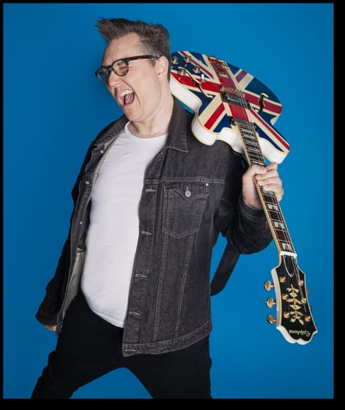 A musician with glasses joyfully poses against a bright blue background, holding a Union Jack guitar.