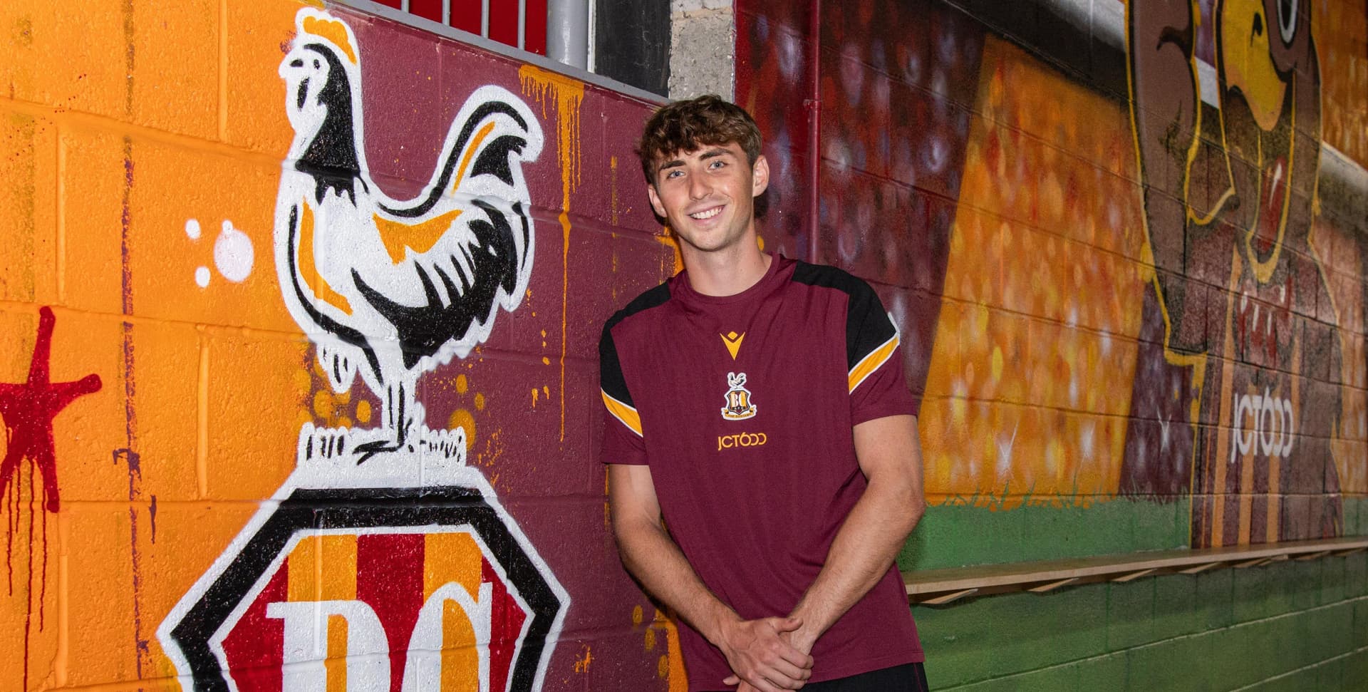 Young man in maroon sports shirt next to vibrant rooster mural