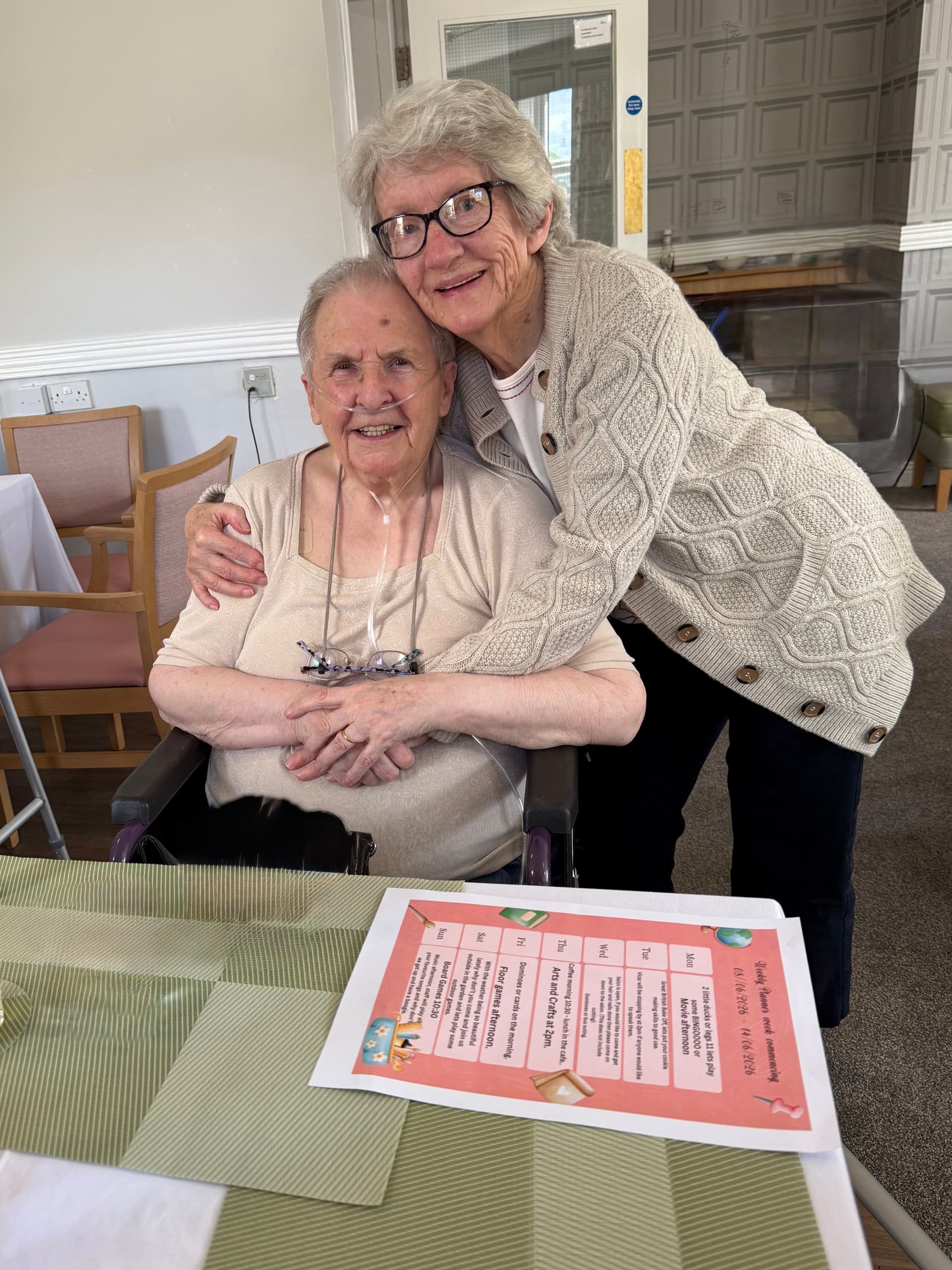 Two elderly women smiling and embracing in a cozy dining room