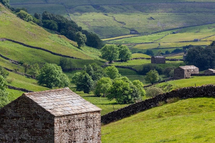 Lush green hills and stone barns dot the landscape of the Yorkshire Dales, framed by dry stone walls and trees.