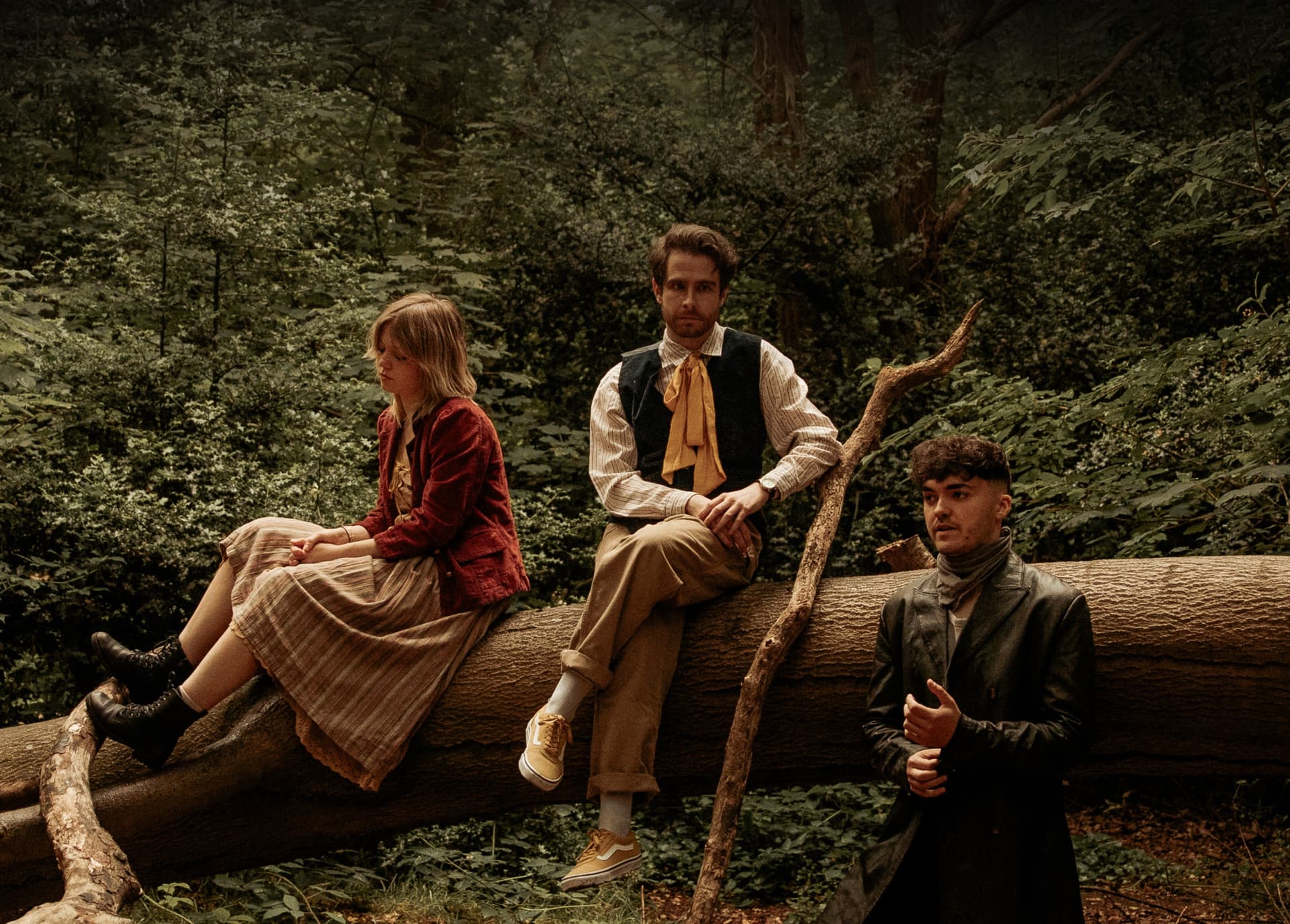 Three people in vintage clothing sitting on a fallen tree in a forest