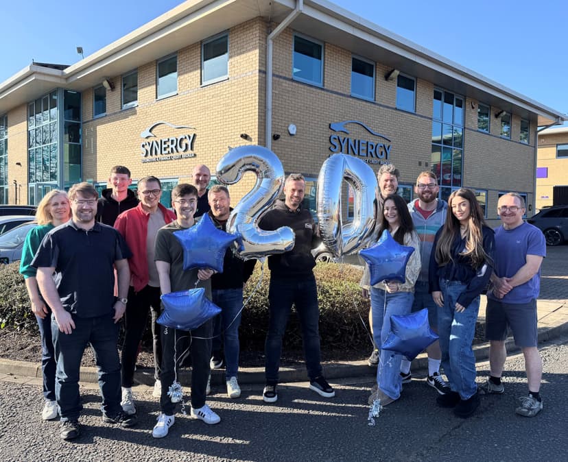 A diverse group of people celebrating outside Synergy's modern building, holding balloons and stars for a 20-year anniversary.