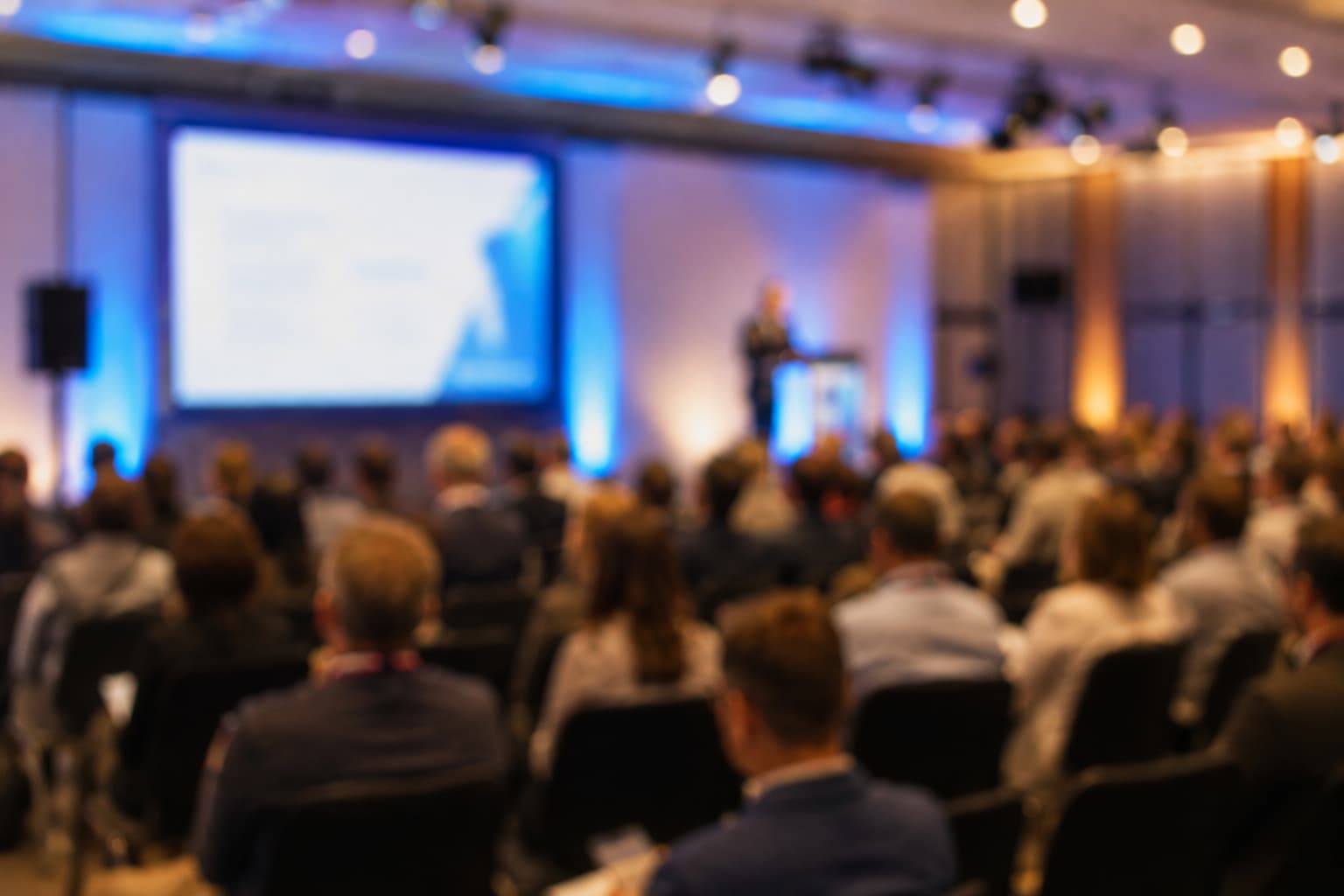 Blurry conference audience with speaker on stage in blue-lit room