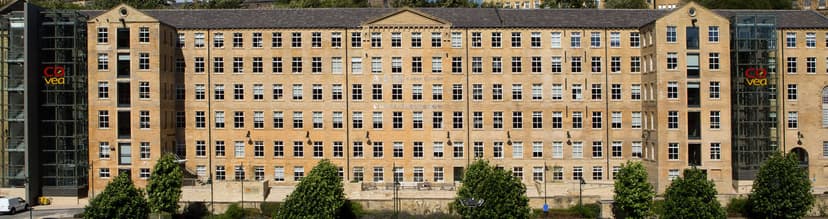 Historic Dean Clough mill with large windows, modern glass extensions, and lush greenery in front, showcasing urban regeneration.