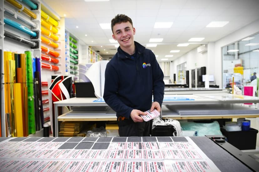 Young man in a workshop smiles while handling printed labels, surrounded by colorful materials and modern equipment.