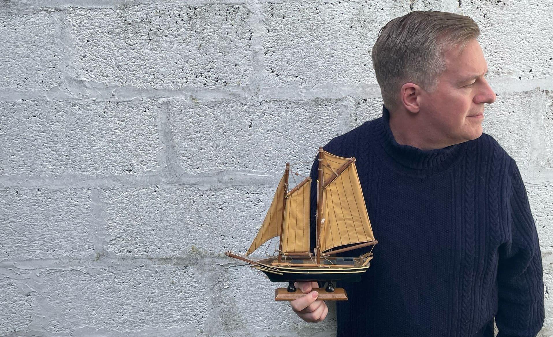 Man in navy jumper holding model sailing ship against stone wall