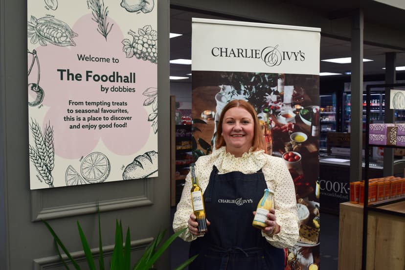Smiling woman in an apron holds rapeseed oil bottles in a bright food hall, surrounded by colorful product displays.