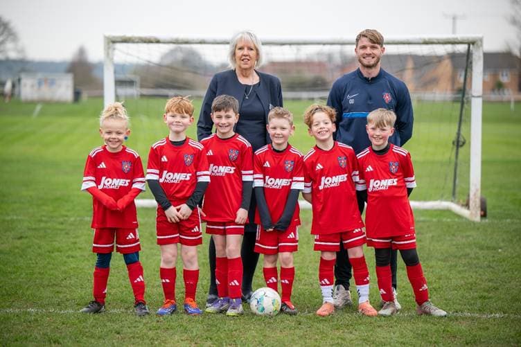 Junior football team in red kits stands on a grassy pitch in Thorpe Willoughby, smiling with their coach and sponsor.