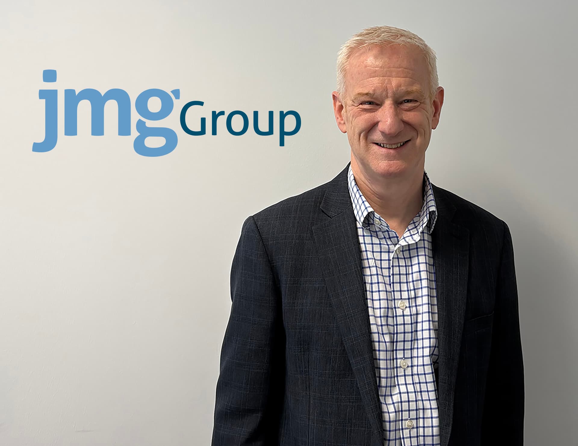 Smiling man in a blazer stands against a light gray wall with "jmg Group" logo, suggesting a professional environment.