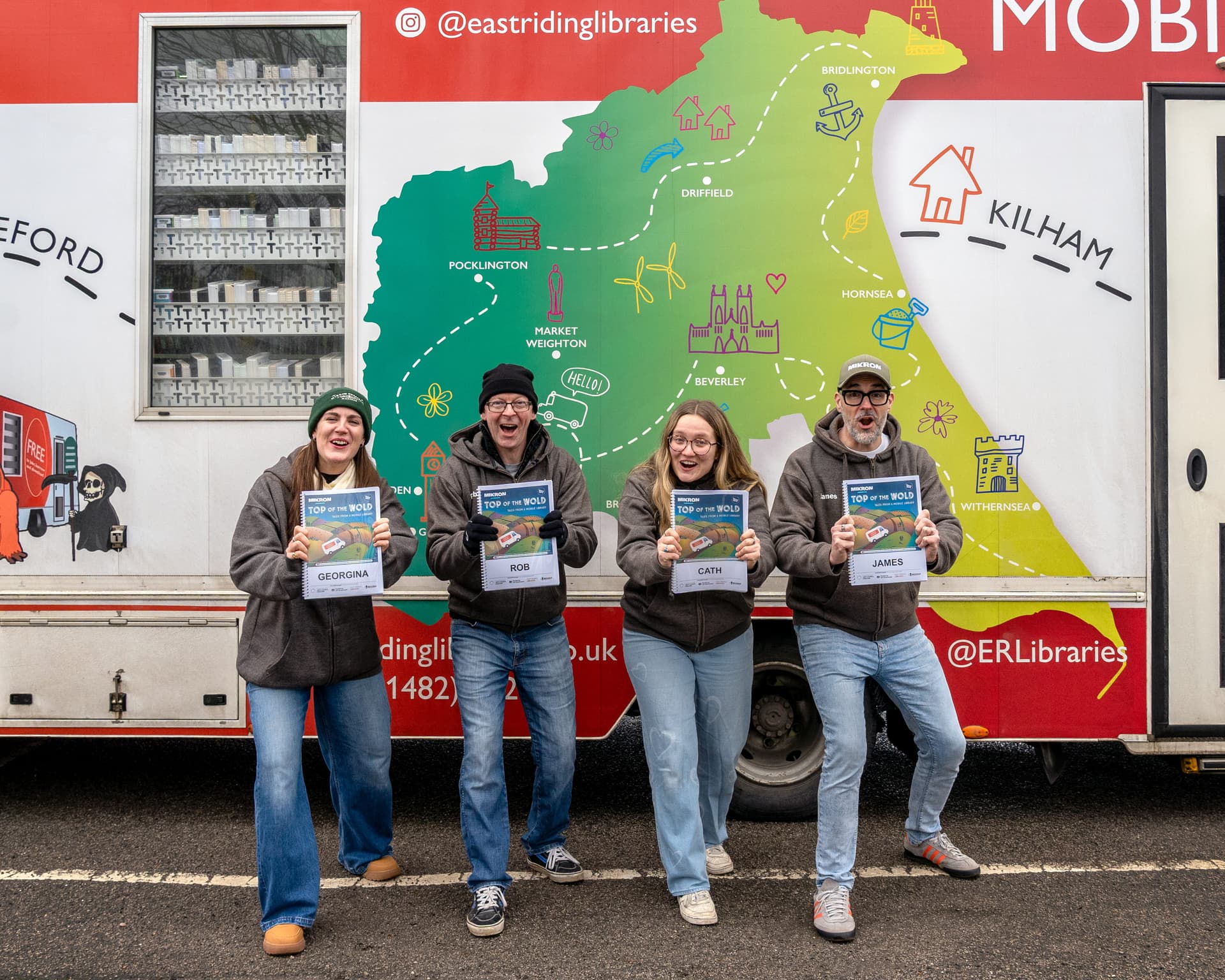 Four cheerful performers hold signs in front of a colorful mobile library truck, set against a vibrant Yorkshire map backdrop.