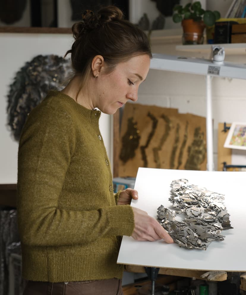 A woman in a cozy studio examines a detailed metallic artwork, surrounded by creative materials and tools.