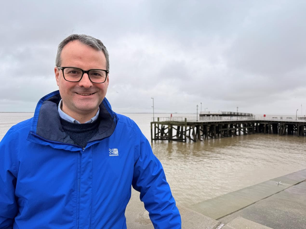 A man in a blue jacket stands by the water, with Victoria Pier in the background under a cloudy sky.