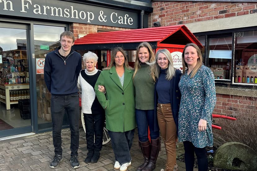 Group of six smiling people outside Pickering Farm Shop Café, with red-roofed kiosk and brick building in the background.