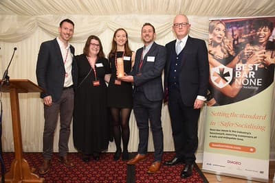 A group of four people in formal attire hold an award, smiling at an event with a banner promoting Best Bar None.