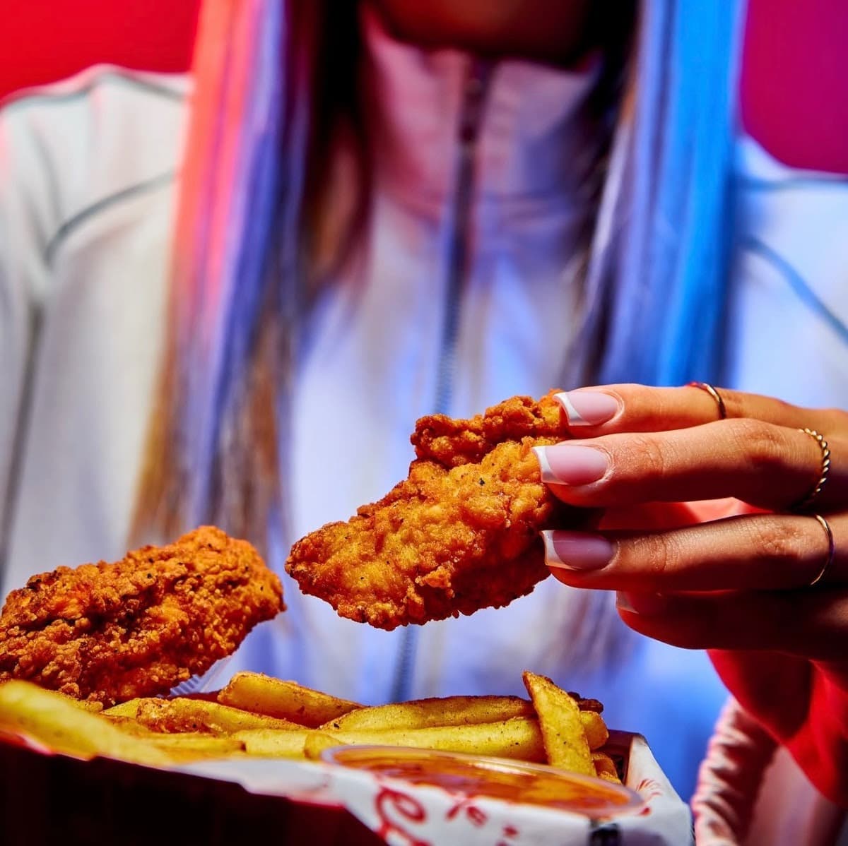 Person holding crispy fried chicken above chips in vibrant lighting