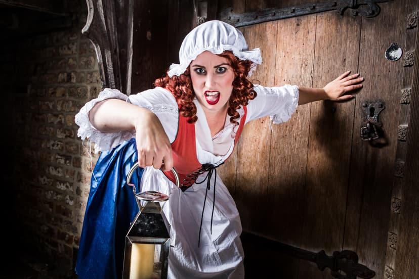 A woman in historical attire, holding a lantern, dramatically poses by a rustic wooden door, evoking a spooky atmosphere.