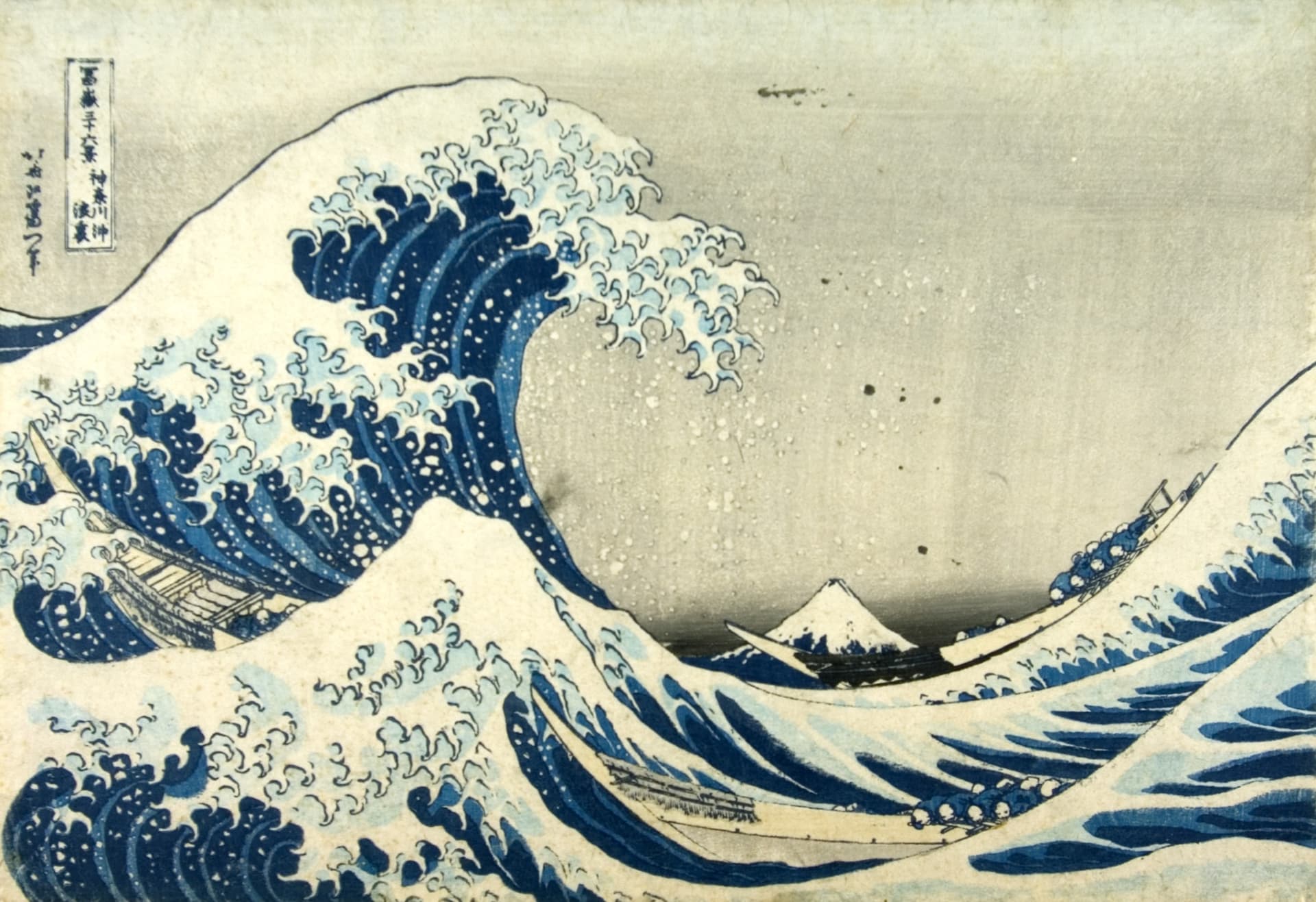 Dynamic blue waves crash against boats, with a distant mountain peak under a cloudy sky, showcasing traditional Japanese art.