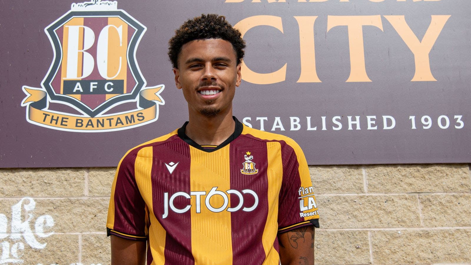 Bradford City AFC player in claret and amber kit standing in front of club sign