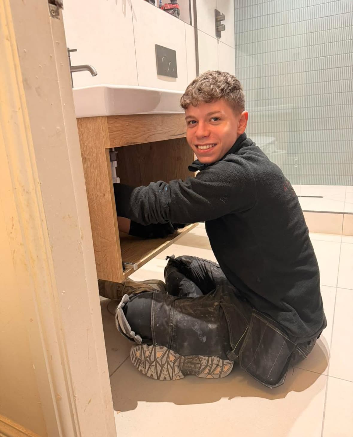 Apprentice plumber working under a sink in a modern bathroom, smiling amidst tools and plumbing fixtures.