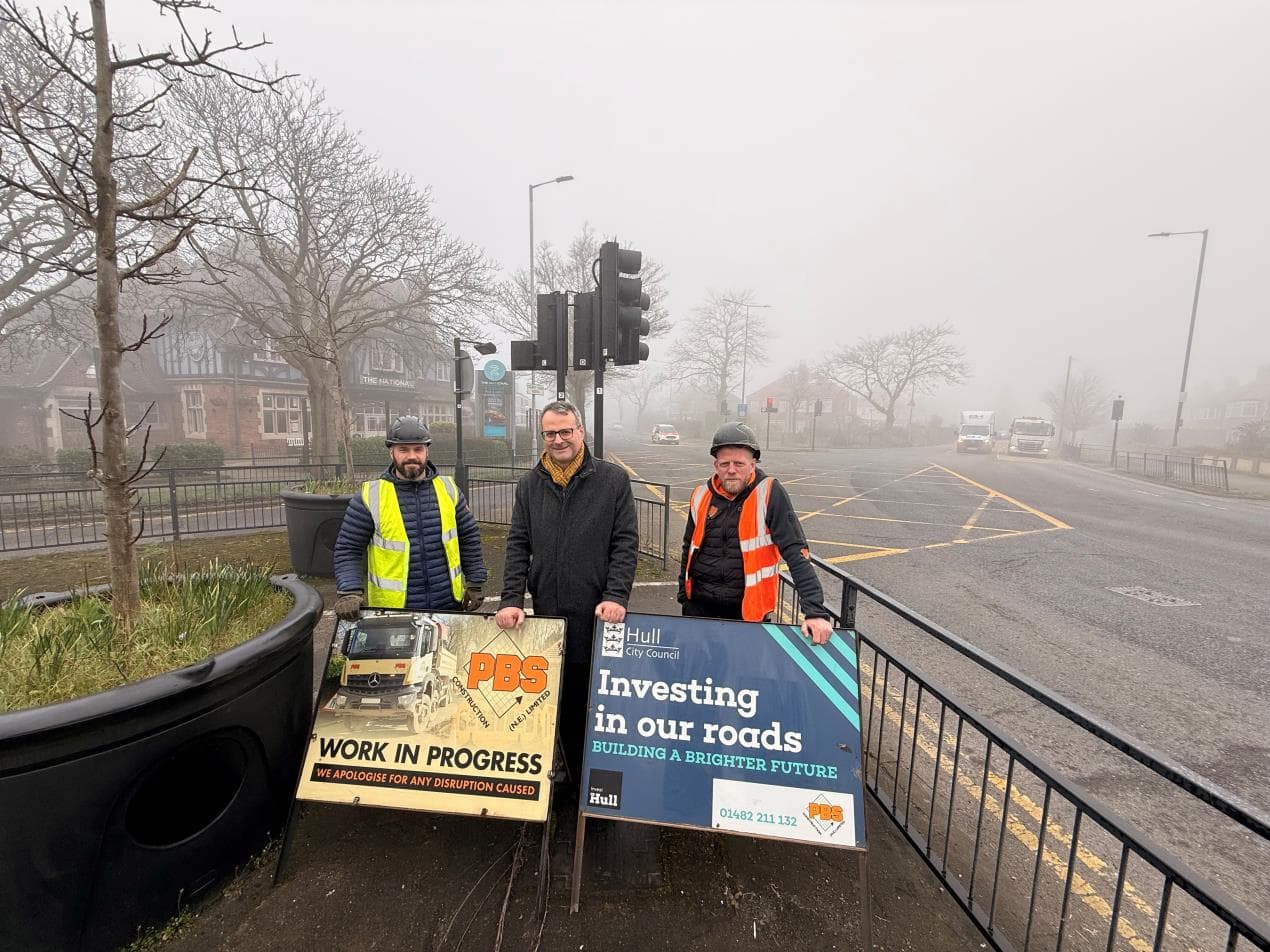 Two workers stand with improvement signs at a foggy intersection in Hull, surrounded by trees and vehicles in the background.