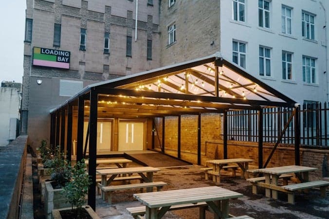 A modern outdoor space with wooden benches and string lights, set against brick buildings in Bradford's Loading Bay.