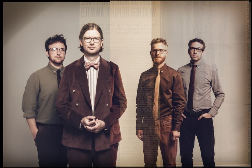 Four musicians pose together, dressed in stylish attire, with a vintage backdrop hinting at a historical atmosphere.
