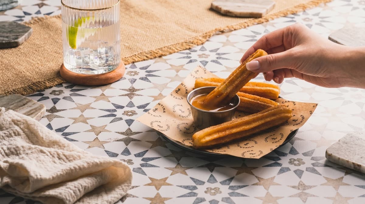 Hand dipping churro into chocolate sauce on patterned table