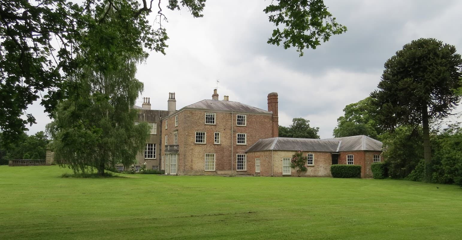 Historic brick estate surrounded by lush green lawn and trees under a cloudy sky in Scampston, Yorkshire.