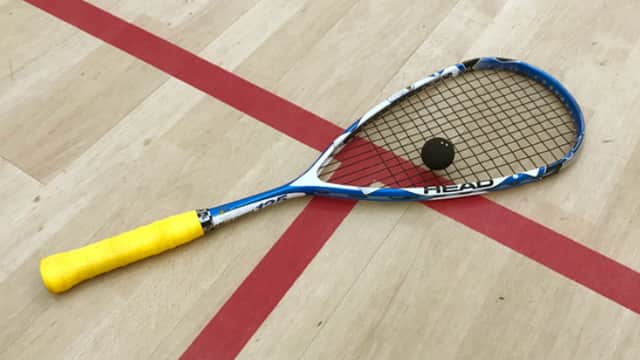 A squash racket with a yellow grip lies on a wooden court beside a black squash ball, marked by red lines.