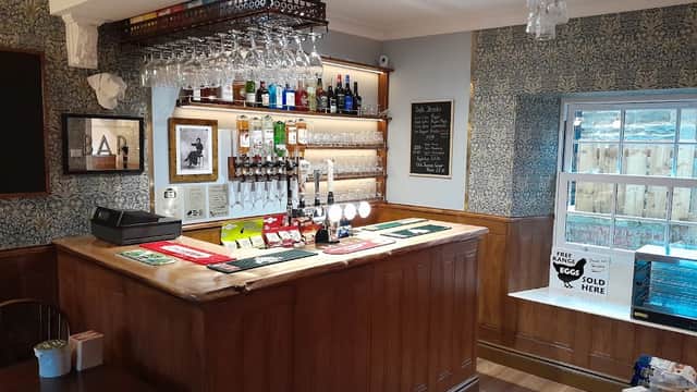 Cozy bar area with wooden counter, shelves of drinks, and a window overlooking a garden. Wallpaper adds character.