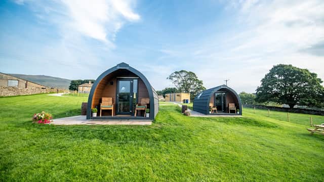 Two glamping pods with wooden decks sit on a grassy hill, surrounded by trees and scenic Yorkshire Dales landscape.