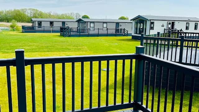 View from a deck overlooking green grass, holiday homes, and a serene lake under a blue sky with scattered clouds.