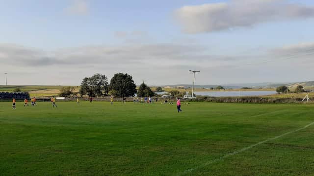 Green football pitch with players in action, surrounded by trees and a scenic landscape under a cloudy sky.