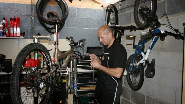 A mechanic repairs a bike in a workshop, surrounded by tools, bike parts, and two mounted bicycles on the wall.