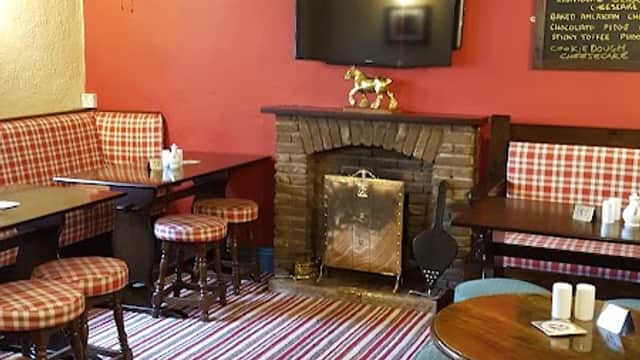 Cozy bar interior with striped carpet, wooden tables, plaid seating, a fireplace, and a TV on the wall.