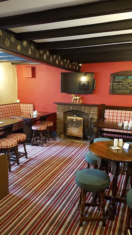 Cozy bar interior with striped carpet, wooden tables, plaid seating, a fireplace, and a TV on the wall.