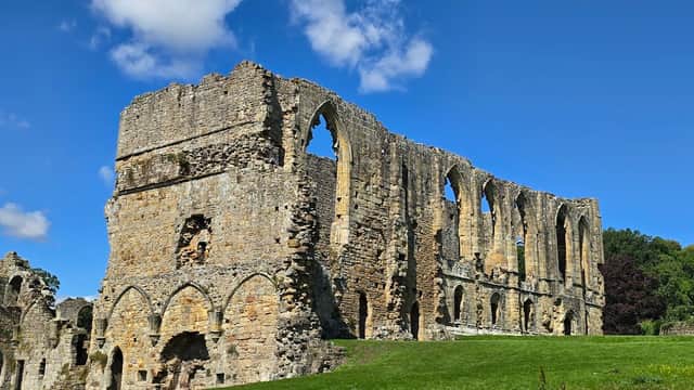Ruins of St Agathaโs Church with arched windows, surrounded by green grass and blue sky in Easby, Yorkshire.