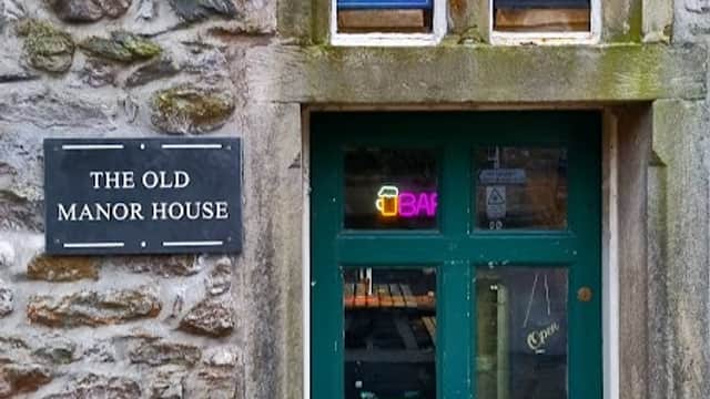 Green door with a sign reading "OPEN" and a plaque that says "THE OLD MANOR HOUSE," surrounded by stone walls.