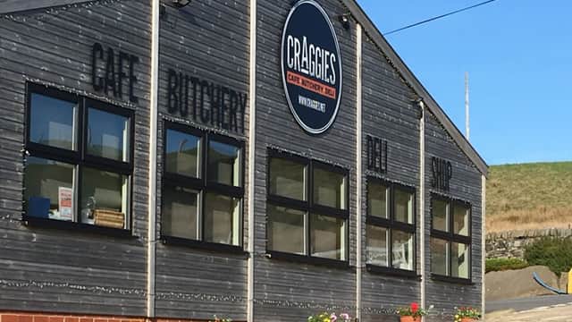Wooden building with large windows, sign reading "Craggies Cafe Butchery Deli Shop," and colorful flower pots outside.