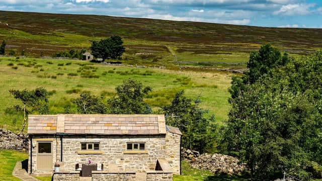 A stone cottage with a slate roof surrounded by green fields and hills under a blue sky with fluffy clouds.