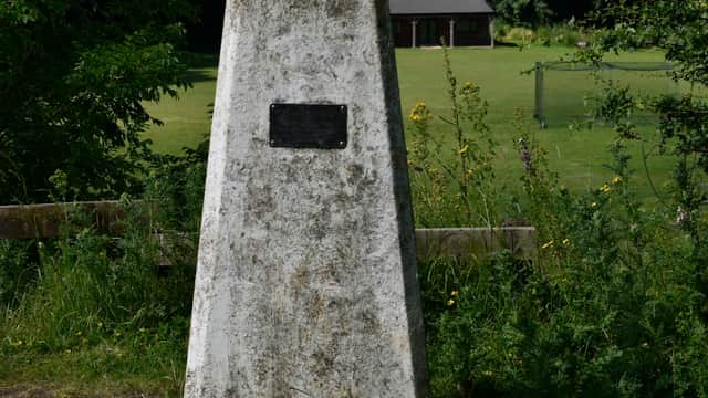 Concrete trig point with a plaque, surrounded by grass and wildflowers, overlooking a field and a wooden building.