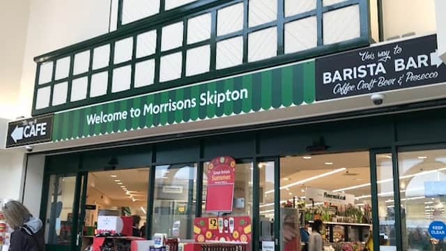 Entrance to Morrisons in Skipton, featuring a clock, café sign, and colorful displays of products outside.