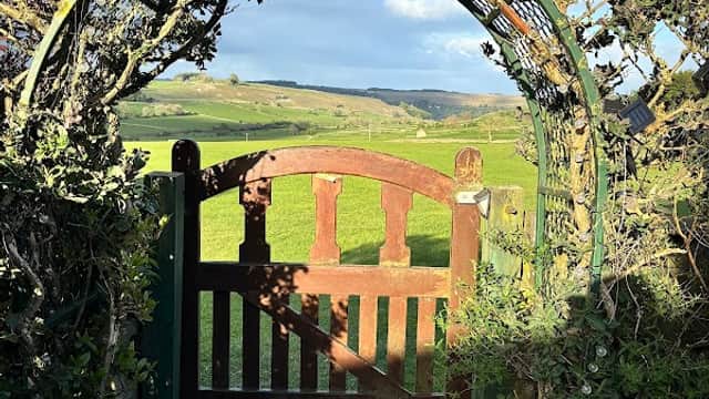 Rustic wooden gate framed by greenery, opening to lush green fields and rolling hills under a clear blue sky.