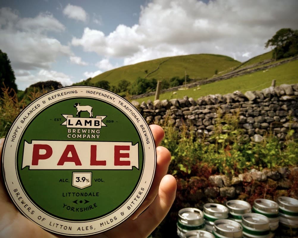A hand holds a beer can labeled "PALE" with green and red design, set against a scenic hillside and stone wall.